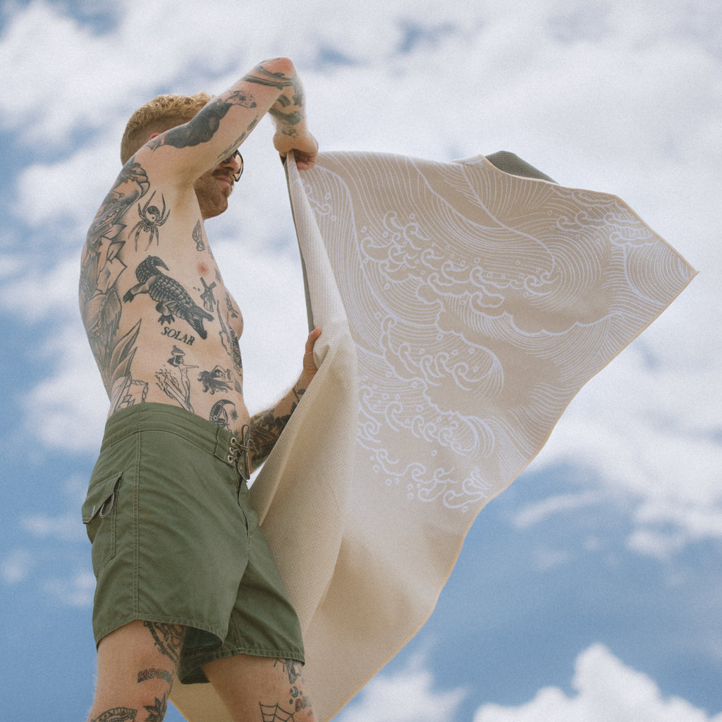 man with tattoos holding beige sand free beach towel with waves design blowing in the wind