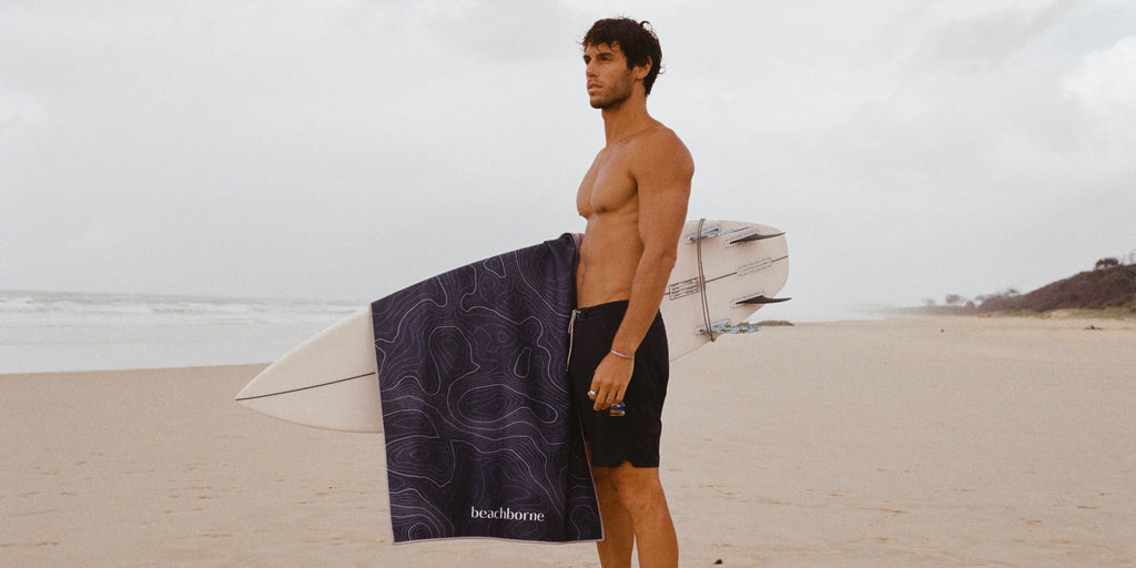 man standing on the beach holding surfboard with black sand free beach towel draped over his surfboard 